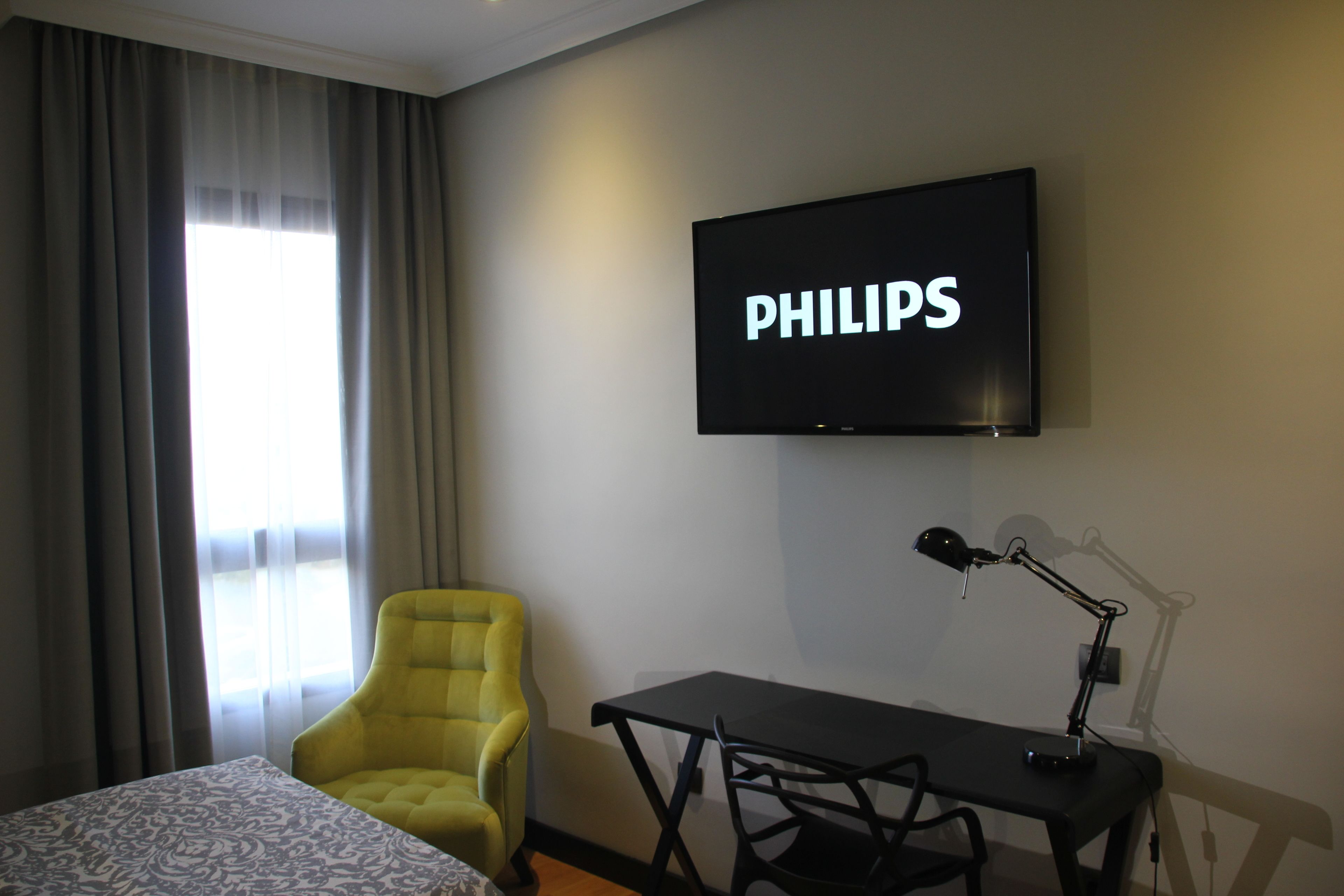 Et hotelværelse med en seng, en gul lænestol, et skrivebord med lampe og et fjernsyn ophængt på væggen, der viser "PHILIPS".