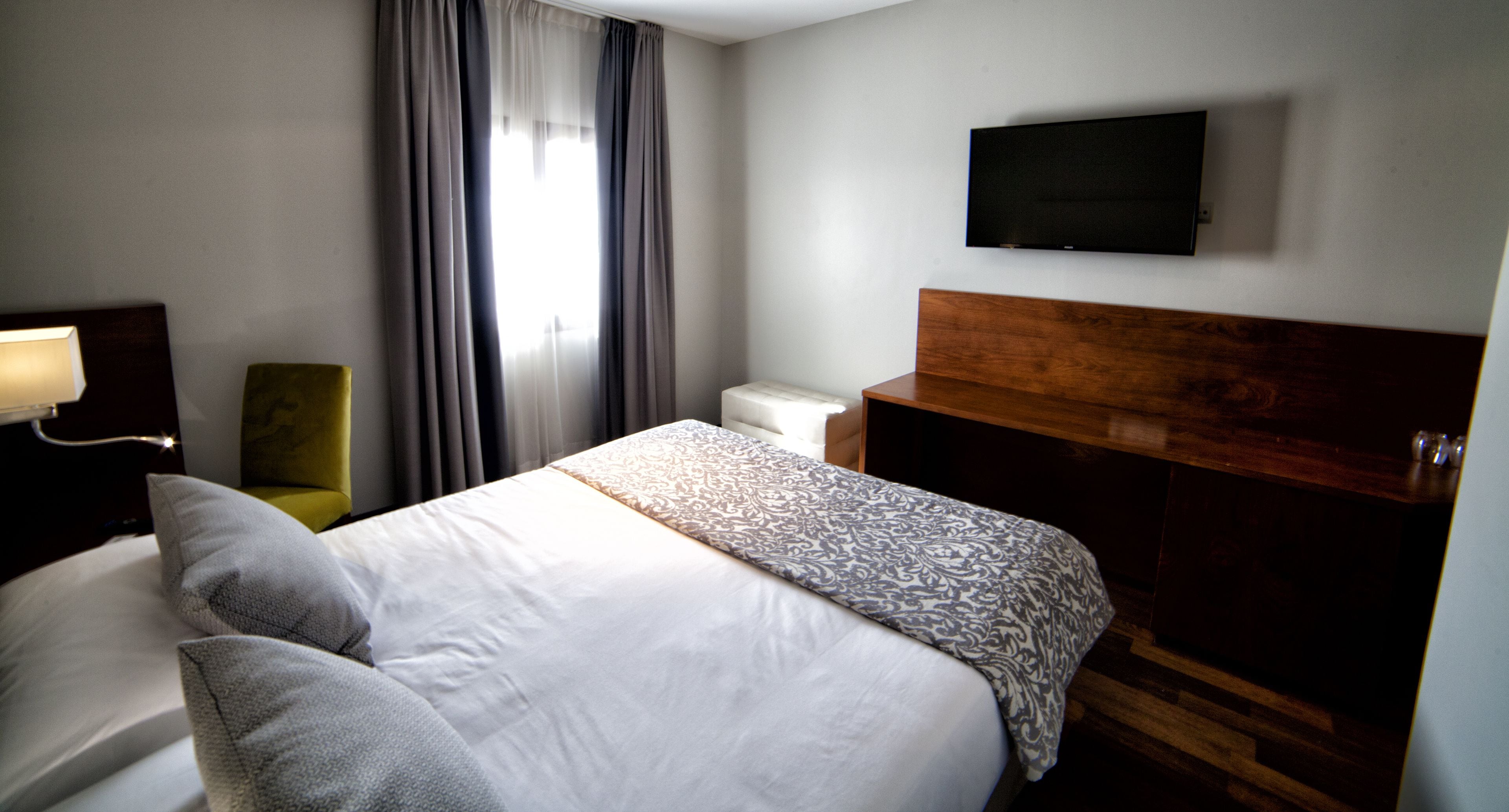 Una habitación de hotel con cama, televisión en la pared, una silla y una ventana con cortinas.
