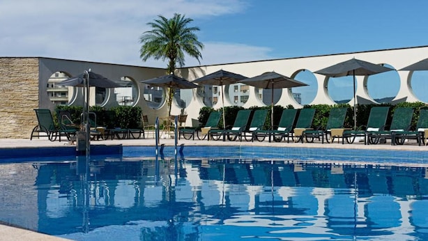 A swimming pool with lounge chairs and umbrellas, surrounded by a modern building with circular windows. - Mendes Plaza Hotel Torre Panorama (Santos)