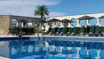Piscina com espreguiçadeiras e guarda-sóis, cercada por um edifício moderno com janelas circulares.