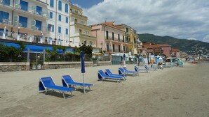 A beach with rows of blue sun loungers, a row of colorful buildings with balconies, and flags on top. - La Balnearia (Alassio)