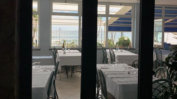 A restaurant with tables and chairs arranged indoors, with large windows offering a view of the outside. - La Balnearia (Alassio)