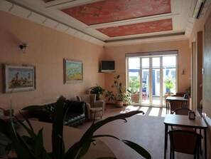 A spacious living area with a sofa, armchairs, a dining table, and potted plants. There is a painting on the wall and a television mounted above it. - La Balnearia (Alassio)