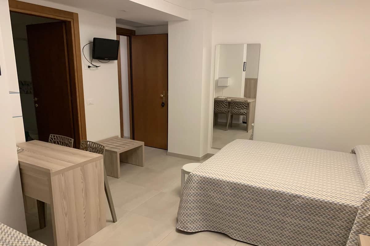 Ein Zimmer mit einem Bett, einem Schreibtisch und einem Sessel. An der Wand befindet sich ein Fernseher und ein Spiegel.