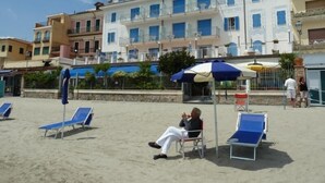 A beach with sun loungers and umbrellas, a building with balconies, and flags in the background. - La Balnearia (Alassio)