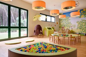 Un salón infantil con piscina de pelotas, ventanas amplias y lámparas colgantes naranjas.