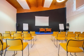 Un salón de conferencias con sillas amarillas, un escenario con una pantalla blanca y dos pizarras.