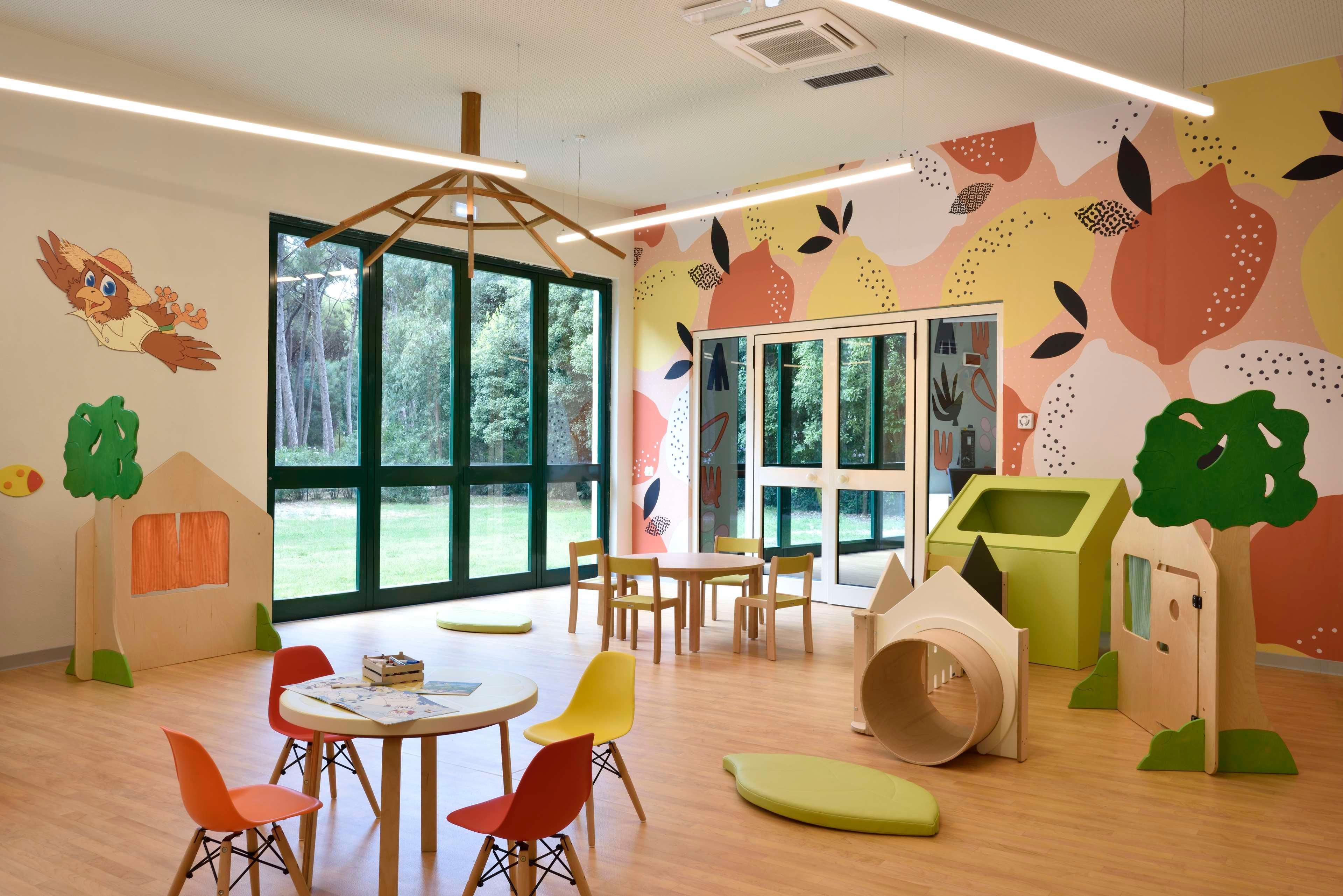 A children's playroom with large windows, wooden play tables, colorful chairs, and playful wall decorations.