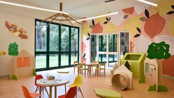 A children's playroom with large windows, wooden play tables, colorful chairs, and playful wall decorations.
