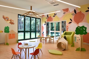 Ein Kinderraum mit großen Fenstern, Holztischen zum Spielen, bunten Stühlen und verspielten Wanddekorationen.