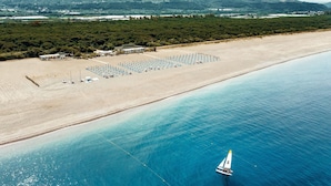 Une vue aérienne d’une plage avec un voilier, une rangée de parasols et un bâtiment doté d’un toit.
