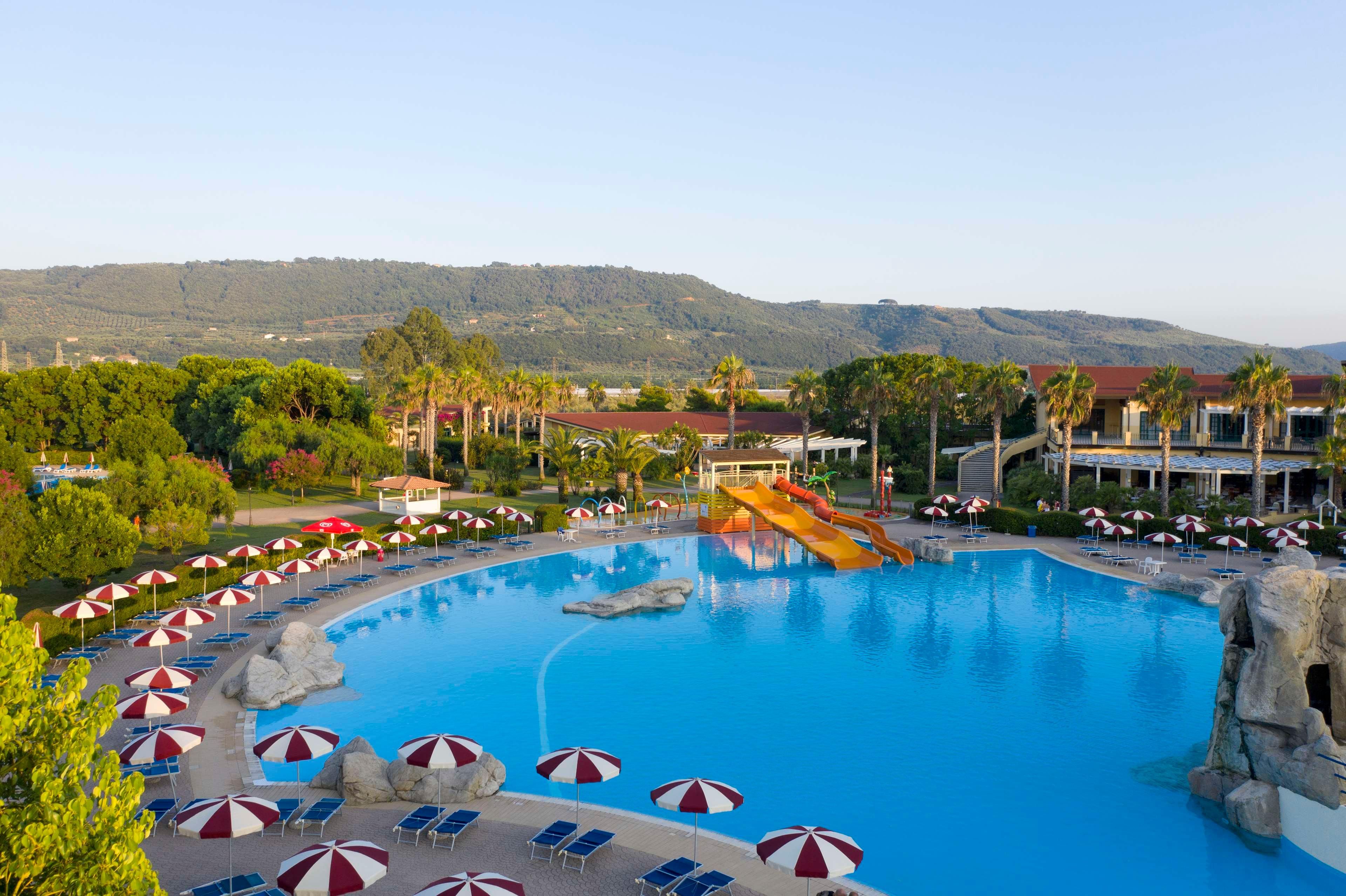 A large swimming pool with a water slide, surrounded by lounge chairs and umbrellas, with a mountainous backdrop.