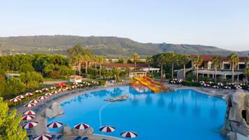A large swimming pool with a water slide, surrounded by lounge chairs and umbrellas, with a mountainous backdrop.