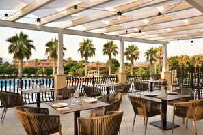 An outdoor dining area with tables and chairs, a pool in the background, and palm trees. - Falkensteiner Club Funimation Garden Calabria (Curinga)