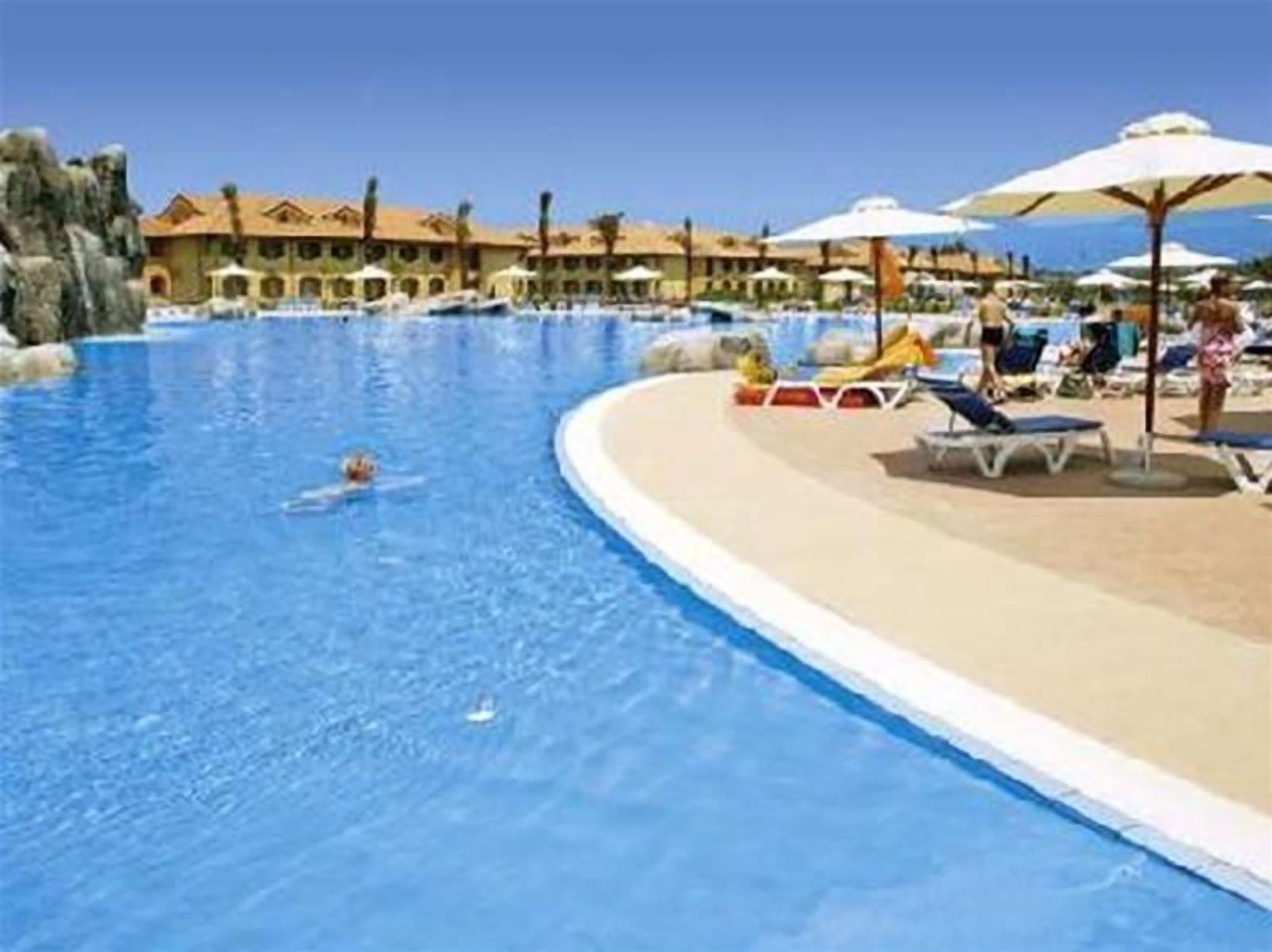Una piscina con sillas de descanso y sombrillas, rodeada de edificios de estilo mediterráneo.
