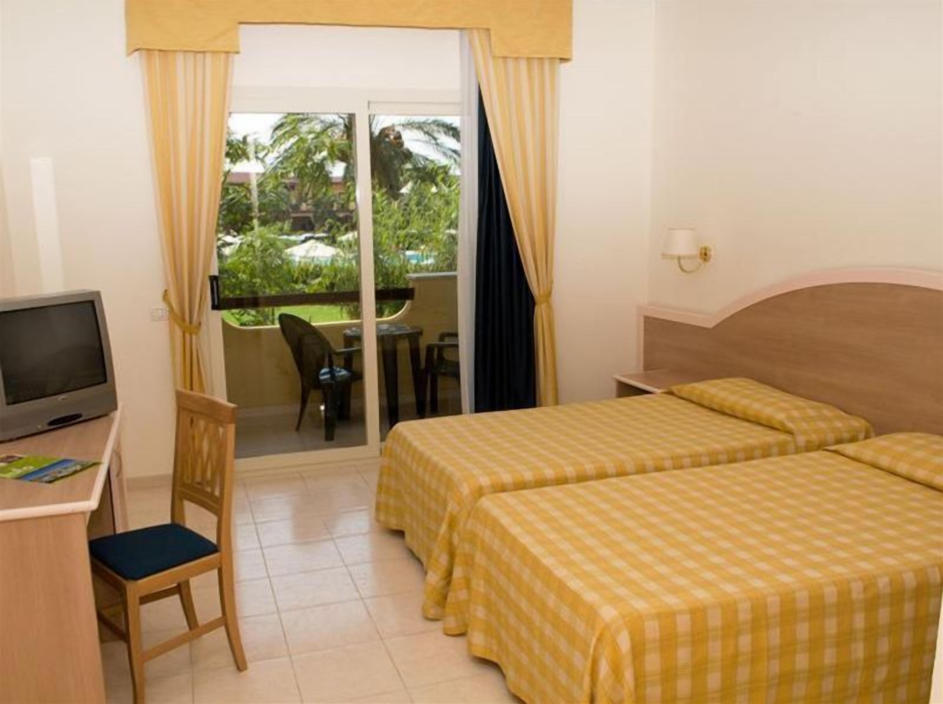 A hotel room with two beds, a desk with a TV, a chair, a balcony with a table and chairs, and a view of greenery and a pool.