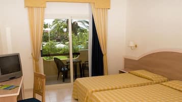 A hotel room with two beds, a desk with a TV, a chair, a balcony with a table and chairs, and a view of greenery and a pool.