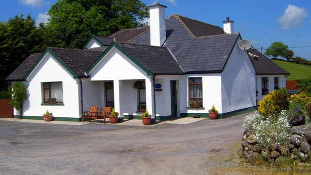 A white building with a green roof and chimney, two windows with flower boxes, and a bench outside. - Valley Lodge (Kilcoman)