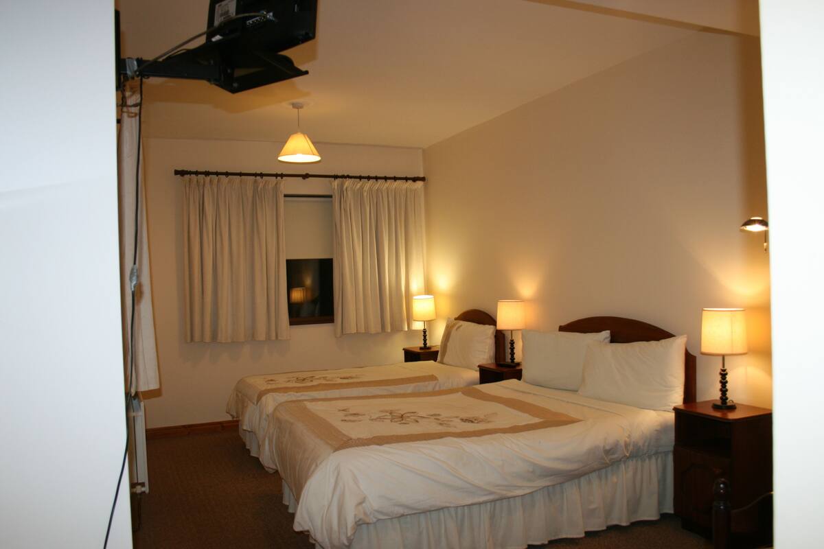 Habitación de hotel con dos camas, una ventana con cortinas, lámparas de noche y un proyector instalado en el techo.