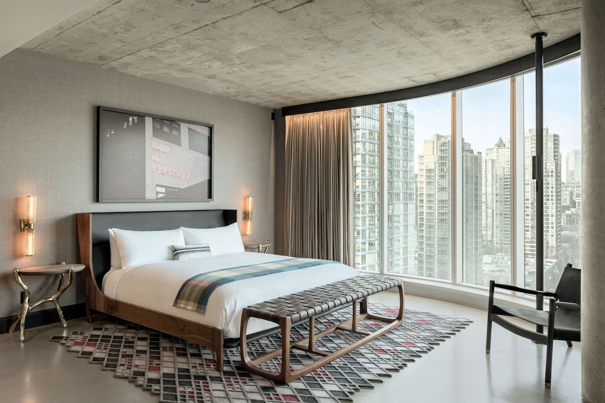 A modern hotel room with a large bed, a bench, a view of the city, and a framed quote on the wall.