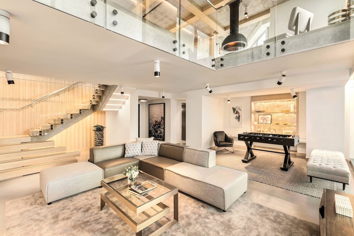 A spacious living area with a modern sofa set, a glass coffee table, and a foosball table. A staircase with glass railings leads to an upper level.