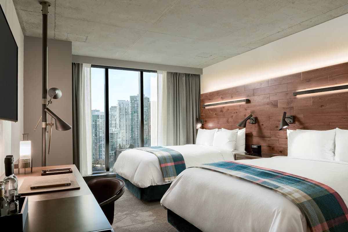 A hotel room with two beds, a desk, a chair, a lamp, and a view of the city.
