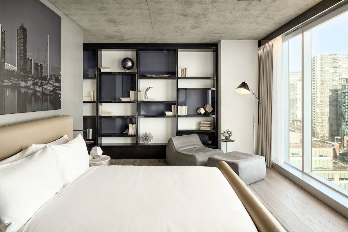 A modern hotel room with a large bed, a built-in bookshelf, a seating area, and a large window with a city view.