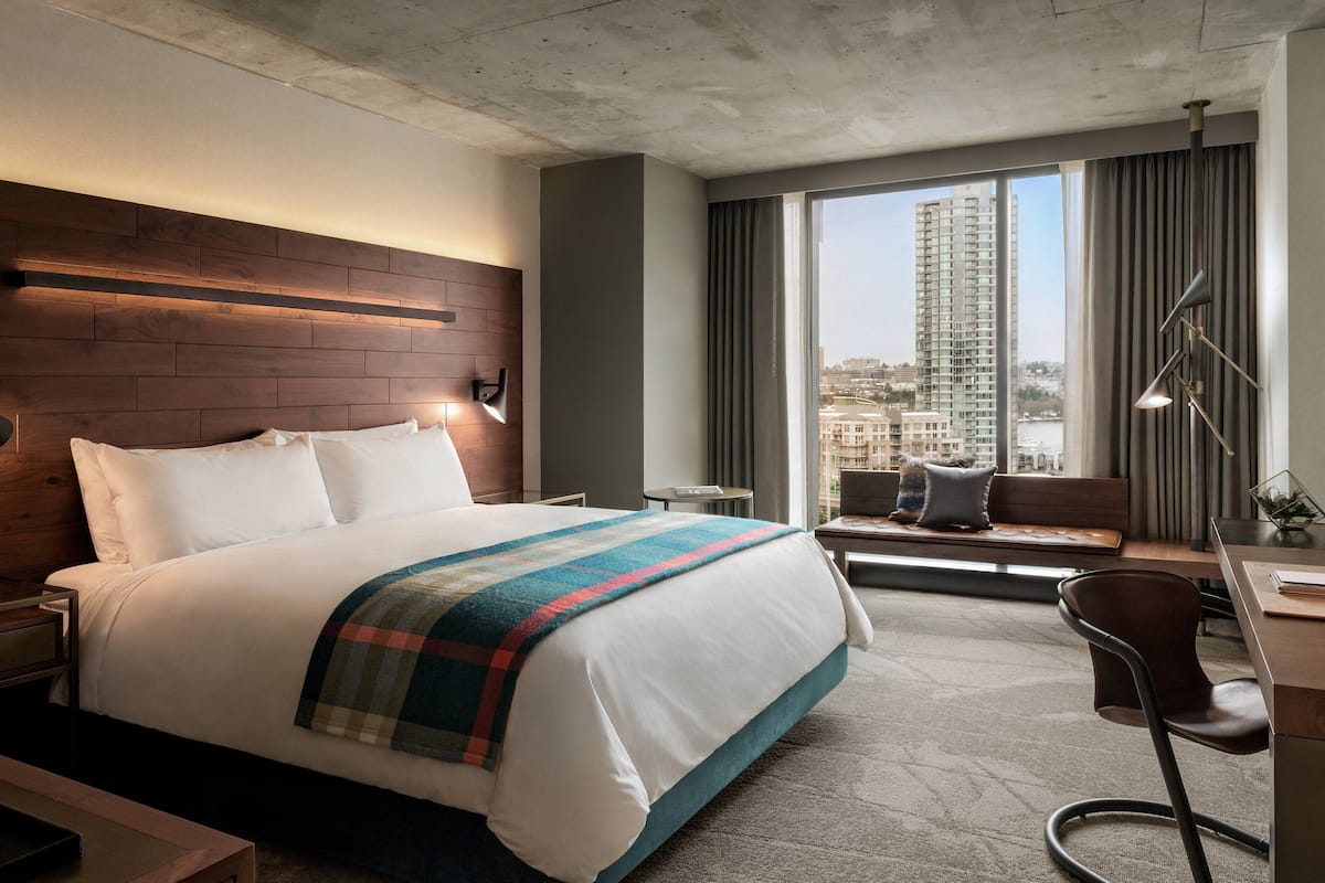A modern hotel room with a large bed, a desk, a chair, and a view of the city through large windows.