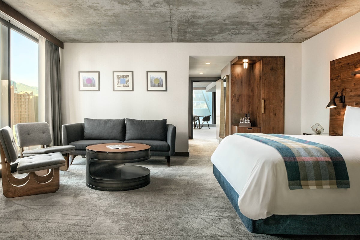 A modern hotel room with a large bed, a grey sofa, a round coffee table, and a unique wooden headboard.
