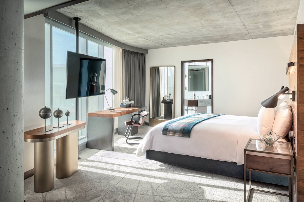 A modern hotel room with a large bed, a desk with a flat-screen TV, a chair, a mirror, and a nightstand.