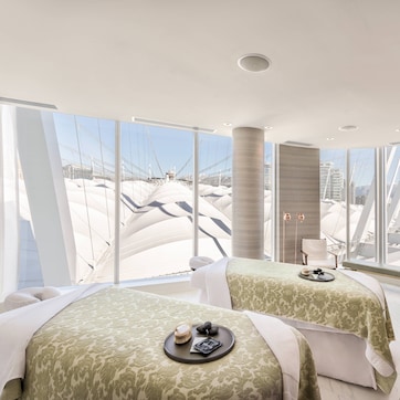 A modern spa room with two massage tables, a small table with a globe and candles, and large windows offering a view of a cityscape.