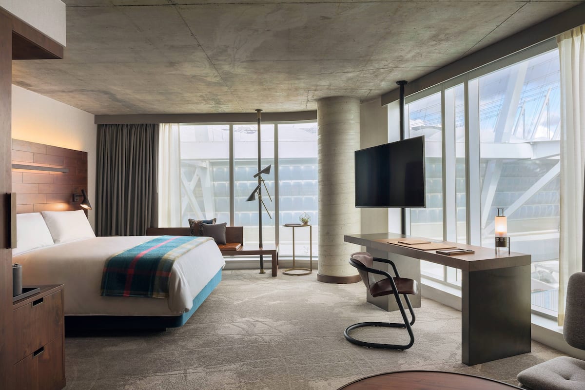 A modern hotel room with a large bed, a desk, a TV, and a view of a bridge through large windows.