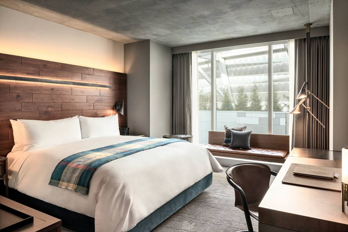 A modern hotel room with a large bed, a desk, a chair, and a window with curtains.