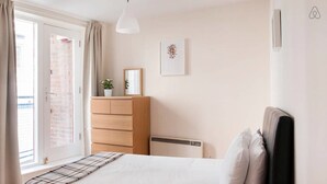 Un dormitorio con cama, cómoda, radiador, ventana con cortinas e imagen colgada en la pared.
