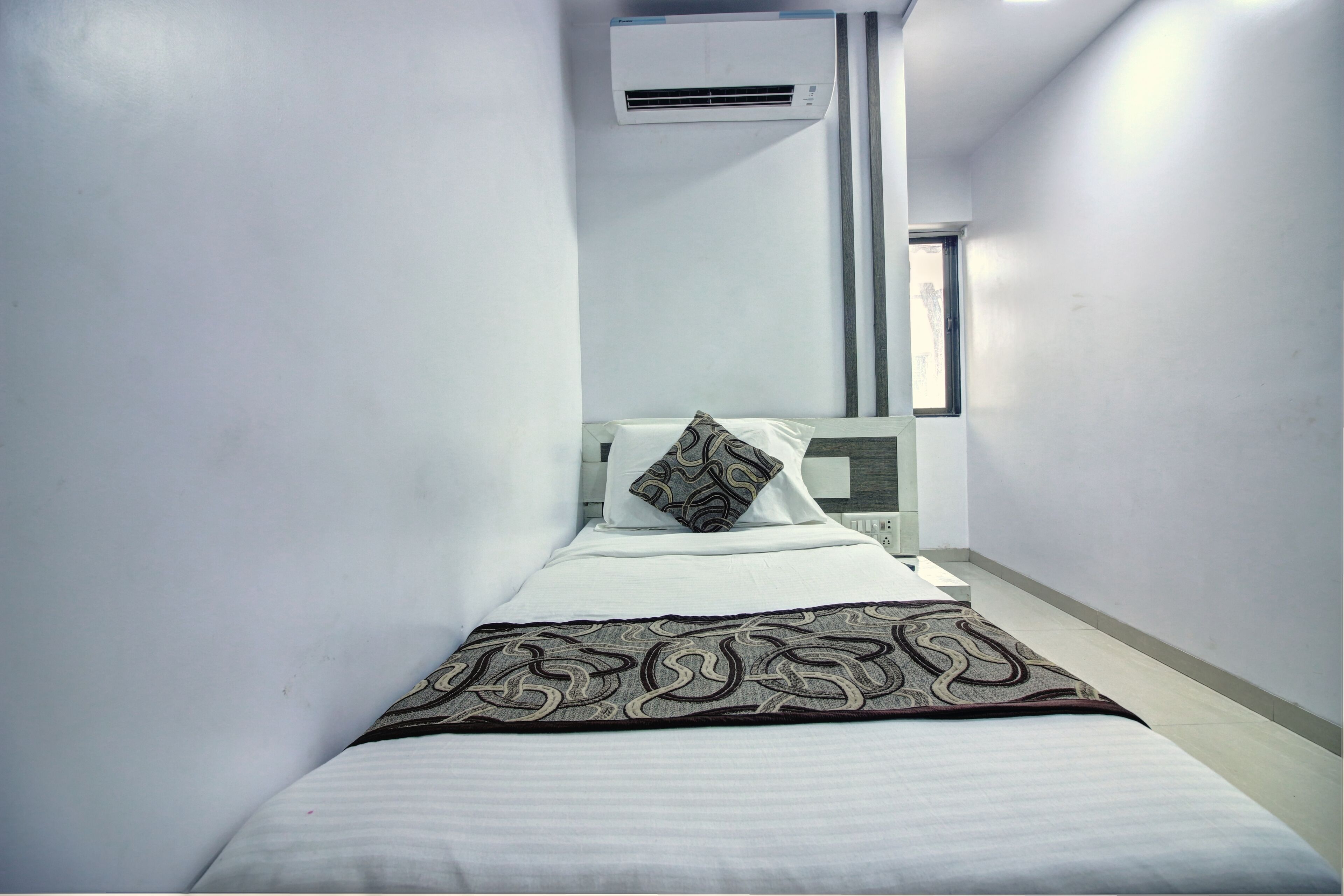 A narrow hotel room with a single bed, a patterned throw blanket, and an air conditioning unit mounted on the wall.