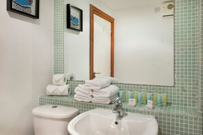 Casa de banho com lavatório, sanita e espelho. Toalhas empilhadas no balcão e artigos de higiene pessoal no lavatório.