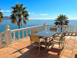 Eine Terrasse mit Tisch und Stühlen, mit Blick auf ein Gewässer und Palmen.