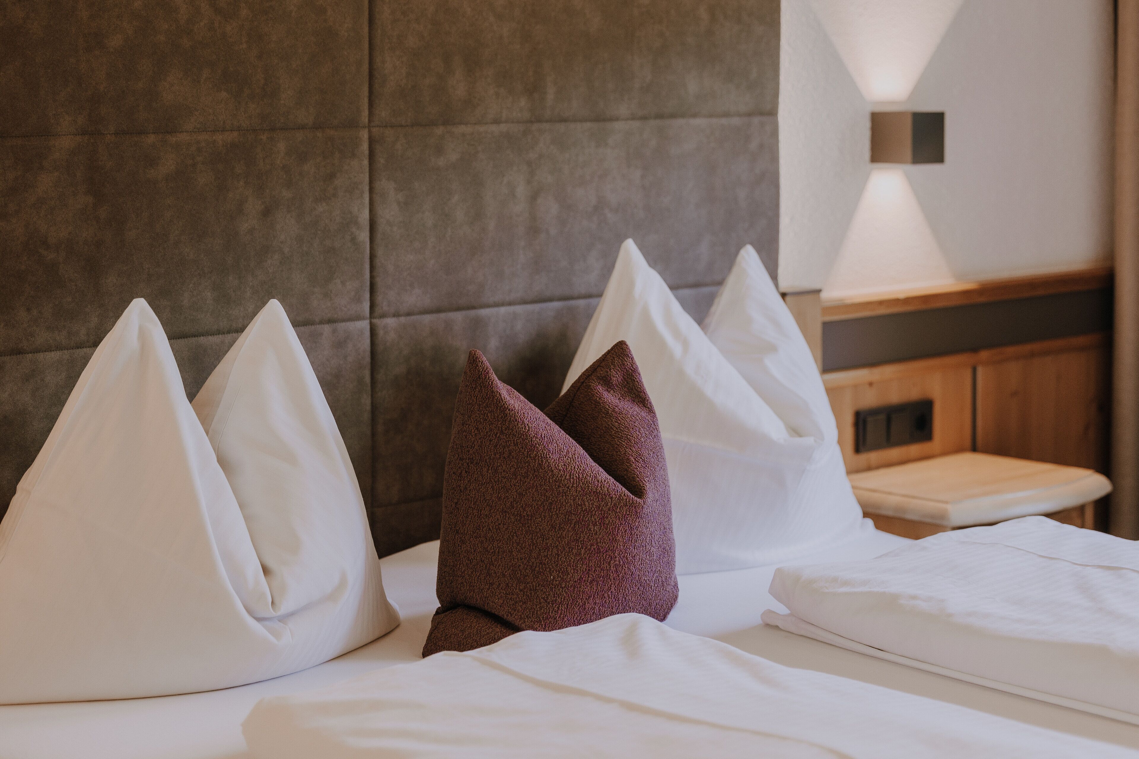 A hotel room with a bed, pillows, a nightstand, and a wall-mounted light fixture.