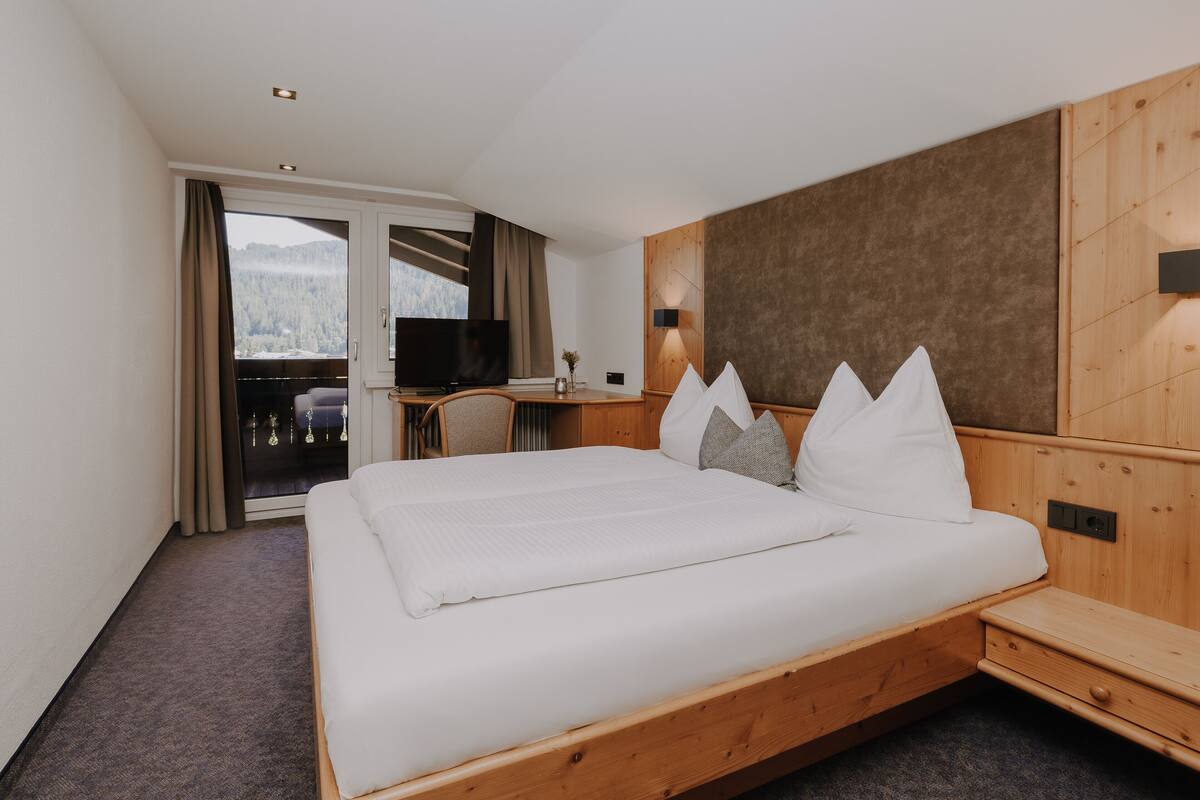 Ein Hotelzimmer mit einem großen Bett, einem Holzkopfteil, einem Schreibtisch mit Stuhl, einem Fernseher und Blick auf eine verschneite Landschaft durch das Fenster.
