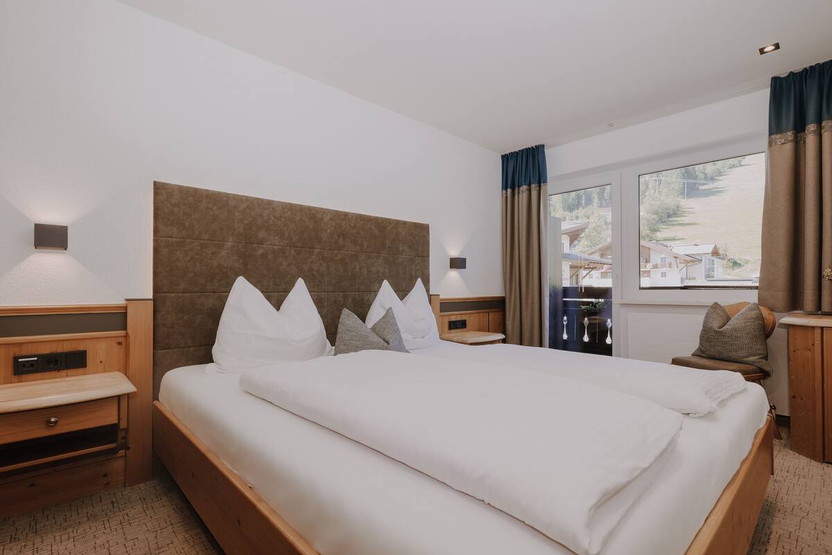 Ein Hotelzimmer mit einem großen Bett, einem Holzkopfteil, Nachttischen und Blick ins Freie durch das Fenster.