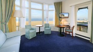 Ett hotellrum med ett stort fönster som vetter mot stranden, en soffa, två fåtöljer, ett litet runt bord och en tv.