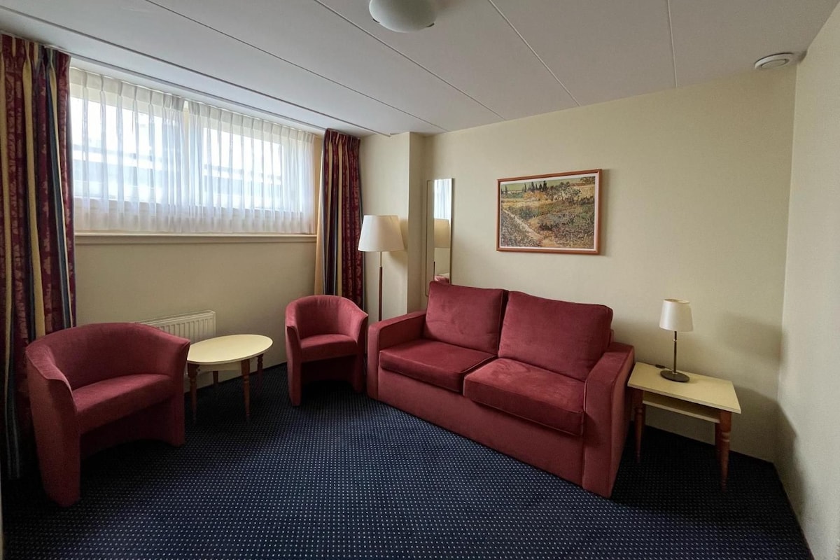 Una habitación de hotel con un sofá rojo, dos sillones, una mesita, una lámpara, un cuadro en la pared y un ventanal con cortinas.