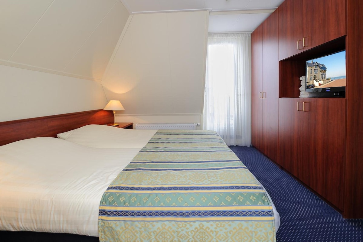 Una habitación de hotel con cama, lámpara de noche, televisor y ventana con cortinas.