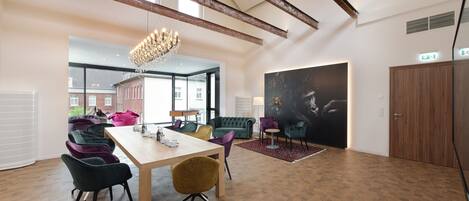 A spacious room with a high ceiling, exposed beams, a chandelier, a large painting, and a view of a building outside.