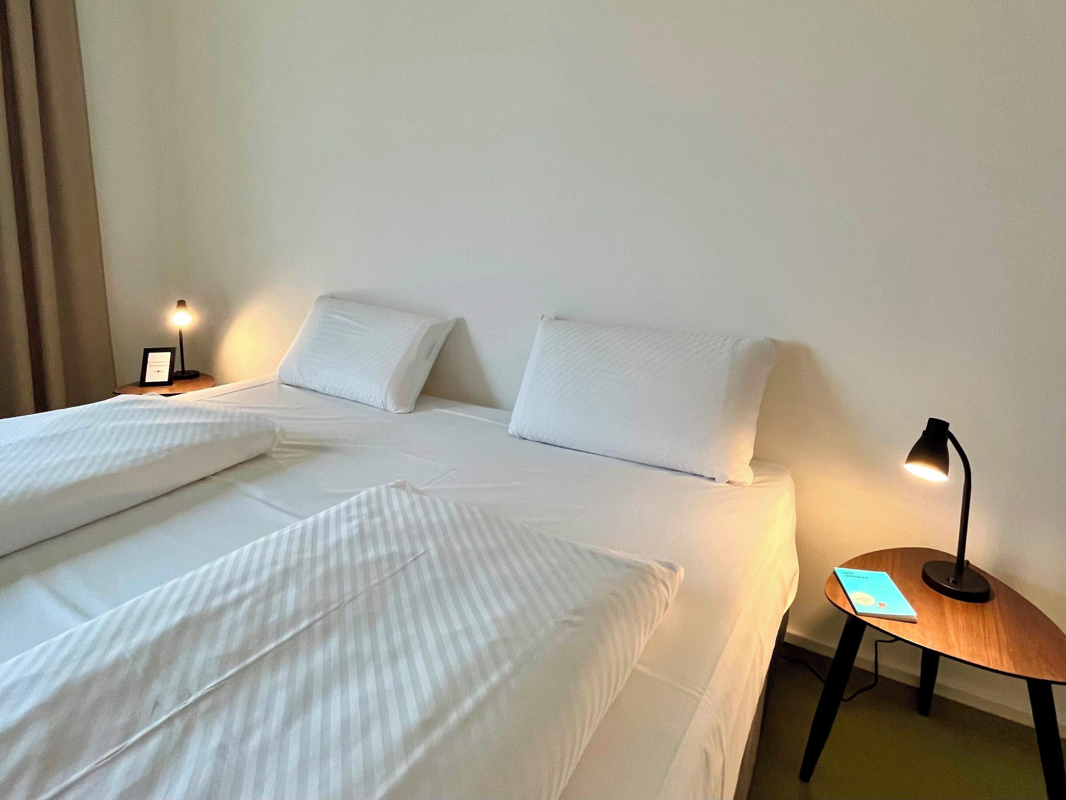 A hotel room with a neatly made bed, two bedside tables with lamps, and a small wooden side table with a book.
