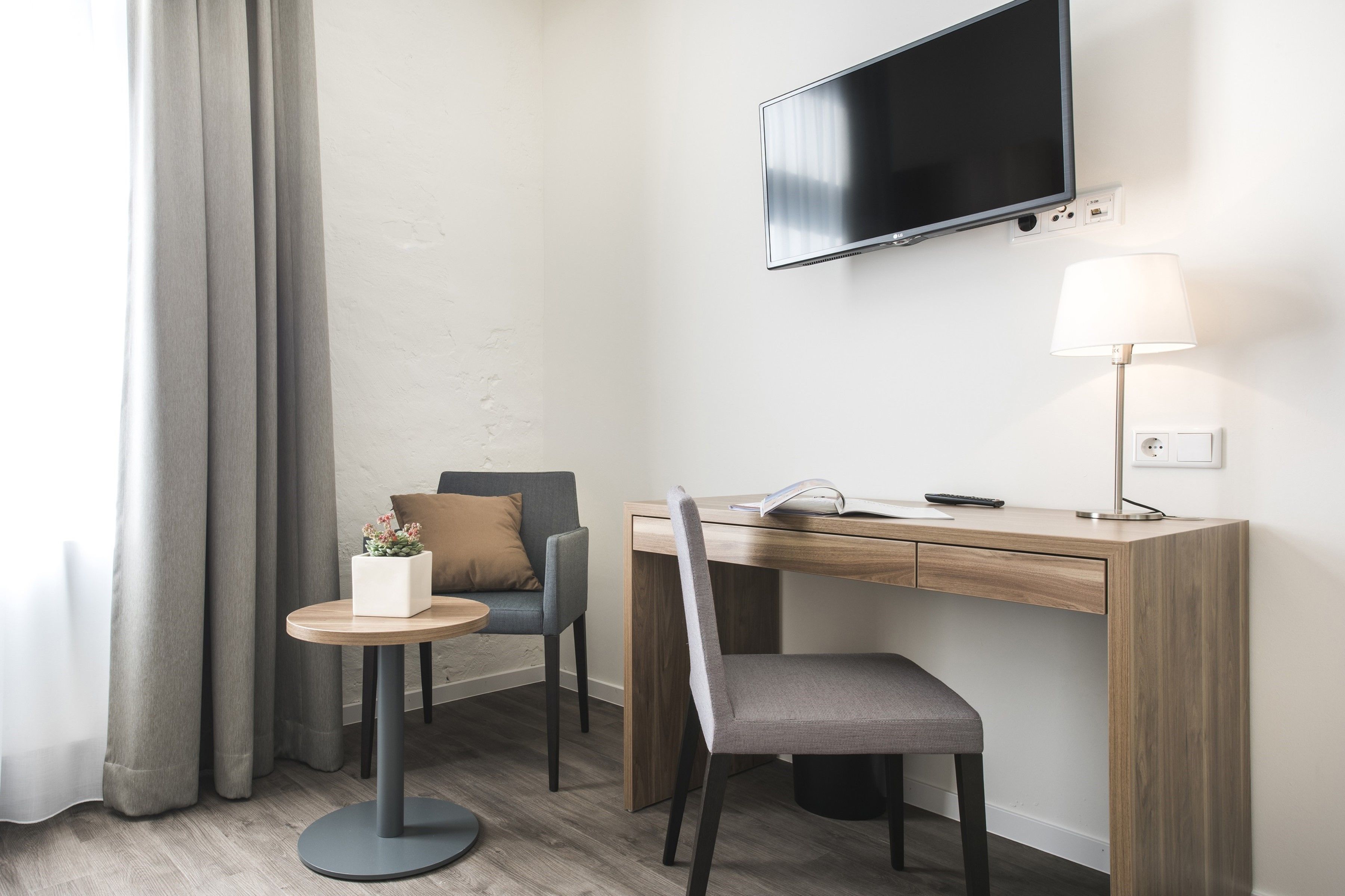 A modern hotel room with a wooden desk, a grey chair, a small round table with a plant, a sofa with a cushion, a TV mounted on the wall, and a lamp on the desk.