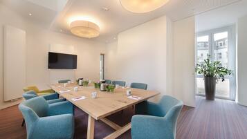 A conference room with a large table, blue chairs, a flat-screen TV, and a potted plant by the window.