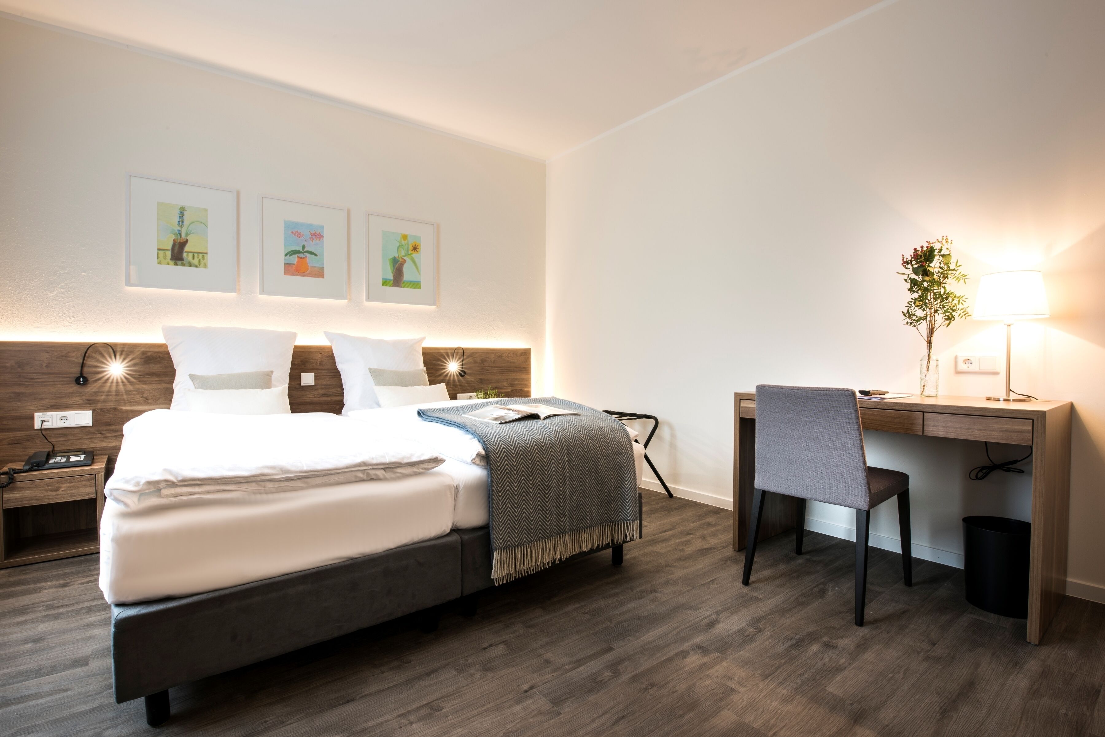 Ein modernes Hotelzimmer mit einem großen Bett, einem Schreibtisch und einem Sessel. Darüber am Bett hängen drei gerahmte Bilder.