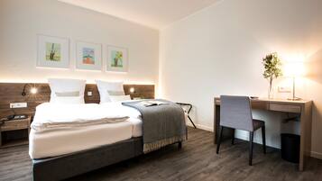 Ein modernes Hotelzimmer mit einem großen Bett, einem Schreibtisch und einem Sessel. Darüber am Bett hängen drei gerahmte Bilder.