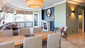 Un salon moderne avec une table à manger en bois, des chaises de couleur beige, un grand luminaire tressé, une cheminée et une vue sur l’extérieur grâce à de grandes fenêtres.